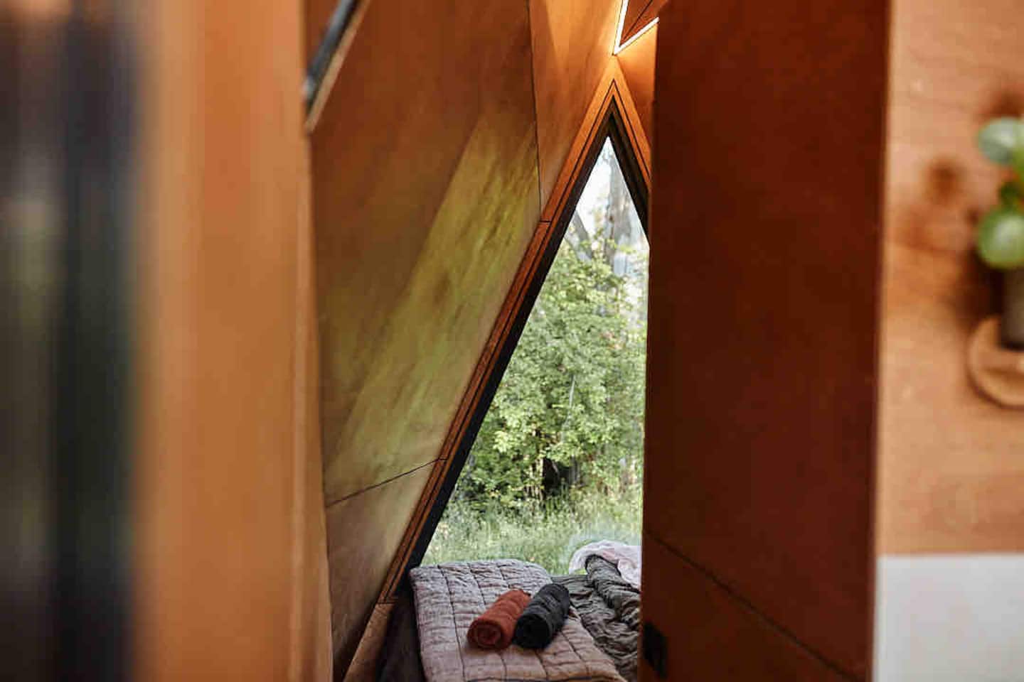 Timber-lined cabin bedroom with a triangular window opening onto the landscape